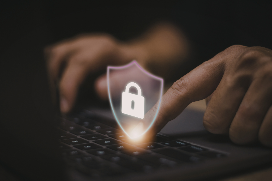 Person typing on a laptop keyboard with a glowing shield and padlock icon symbolising data protection and cybersecurity.